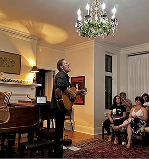 A singer-songwriter performing with acoustic guitar for a seated audience in the Deanery parlor