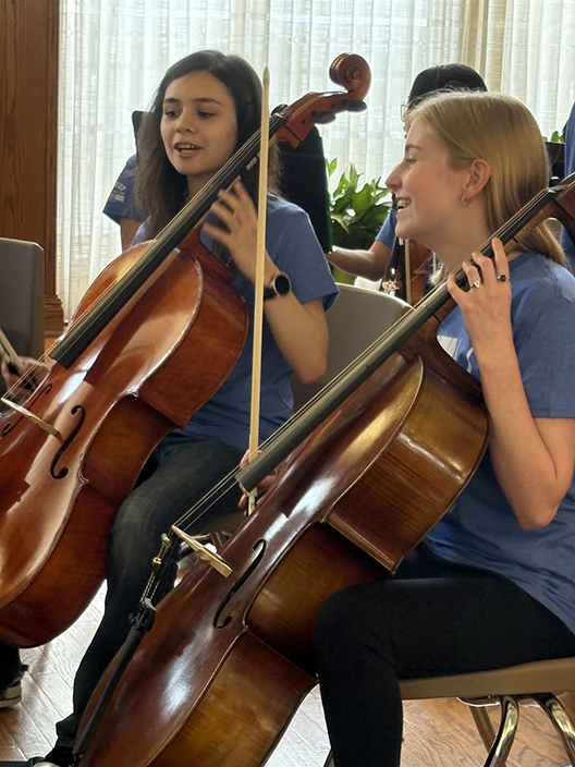 Two young cellists smiling as they play together at The Deanery